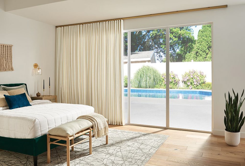 Cozy bedroom with a bed, layered pillows, and a bench at the foot, featuring cream curtains covering sliding glass doors that open to a backyard pool and greenery.