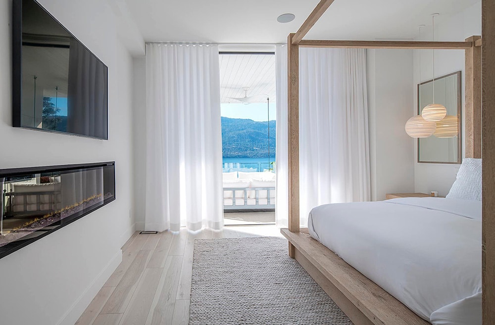 Minimalist bedroom with a wooden canopy bed, white bedding, wall-mounted TV, modern fireplace, and sheer white curtains framing a balcony view of mountains and water.