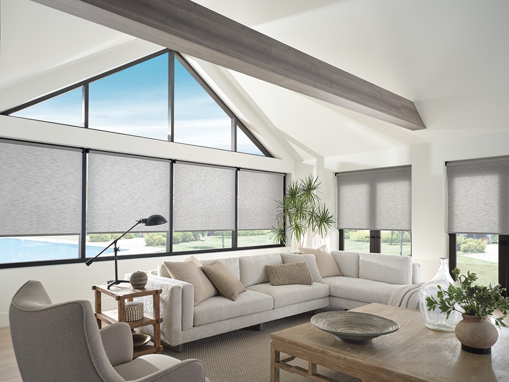 living room with large windows covered by modern roller solar shades, featuring a neutral sectional sofa, wooden coffee table, indoor plants, and vaulted ceiling with exposed beam design.