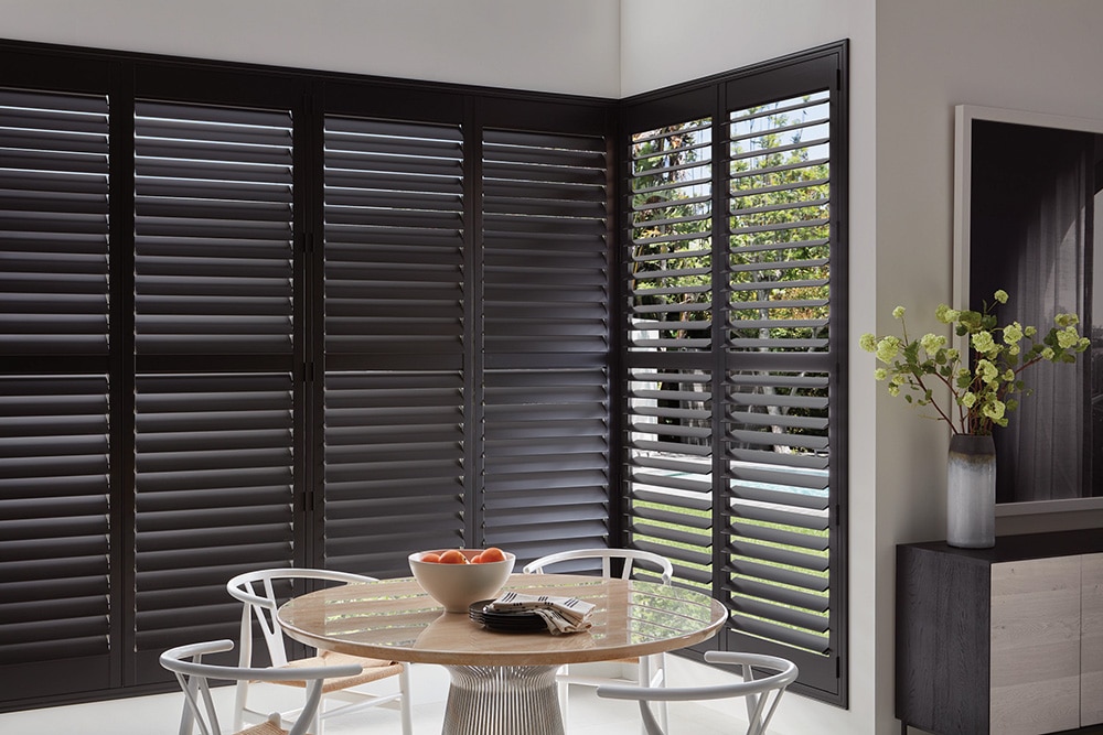 Modern dining area with black plantation shutters on large corner windows, round wooden table with white chairs, and decorative vase with green branches.