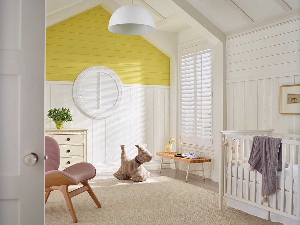 White composite shutters filtering in natrual light into a yellow and white painted nursery.