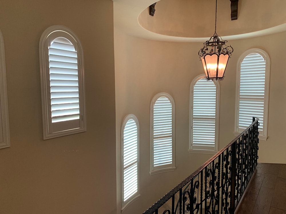 Stairwell with multiple tall arched windows fitted with white plantation shutters, featuring an ornate wrought iron railing and hanging lantern-style light fixture.