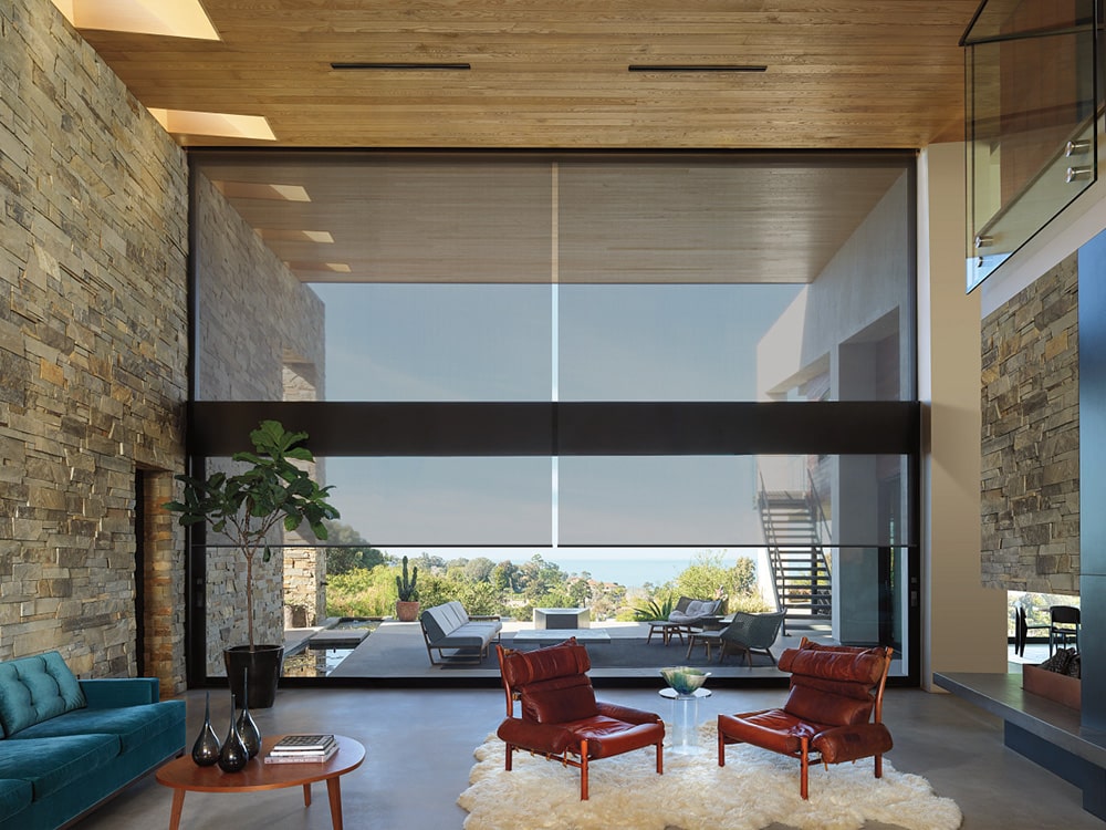 Modern living room featuring large floor-to-ceiling windows with solar shades, stone accent walls, wood ceiling, and stylish furniture including leather chairs, teal sofa, and coffee table, overlooking an outdoor patio and scenic view.